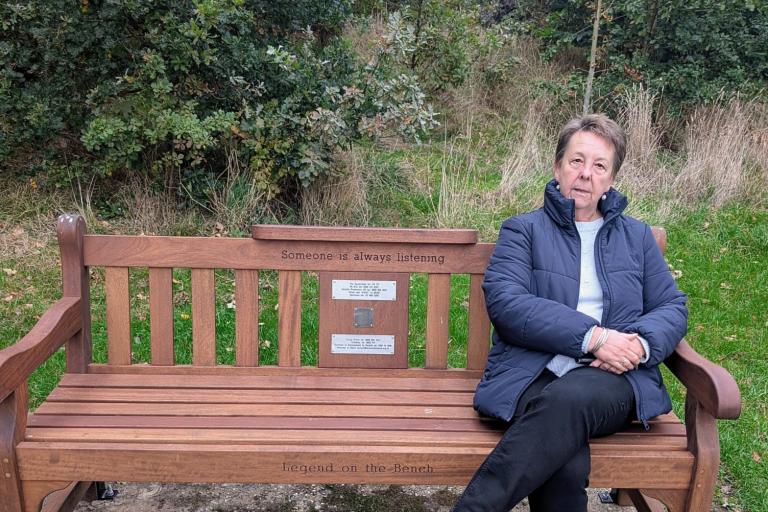 Woman in a blue jacket and dark trousers sitting on a wooden park bench 