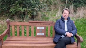 Woman in a blue jacket and dark trousers sitting on a wooden park bench 